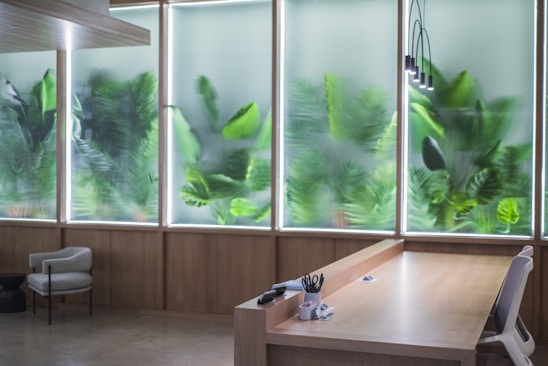 A modern and minimalistic office space featuring a wooden desk and a grey office chair. Large windows with frosted glass create an airy atmosphere, with green leafy plant patterns visible on the glass, giving the illusion of a garden outside. The room is lit softly by pendant lights hanging from the ceiling, and office supplies neatly placed on the desk.