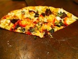 A slice of pizza topped with various ingredients including olives, red peppers, cheese, and fresh herbs is placed on a wooden surface. The crust is golden brown, and the toppings are vibrant, with a mix of green, black, and red colors.