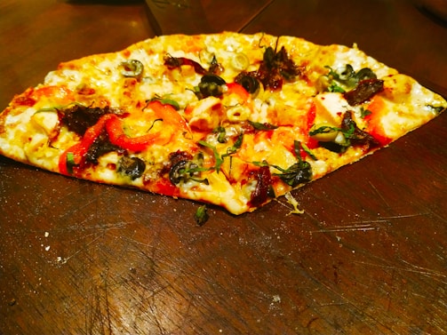 A slice of pizza topped with various ingredients including olives, red peppers, cheese, and fresh herbs is placed on a wooden surface. The crust is golden brown, and the toppings are vibrant, with a mix of green, black, and red colors.