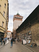 A narrow cobblestone street lined with historic buildings and a medieval tower in the background. The wall on the right is adorned with numerous framed artworks for sale. People are casually walking, some admiring the art, creating a lively yet relaxed atmosphere.