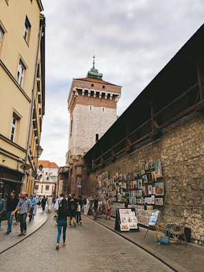 A narrow cobblestone street lined with historic buildings and a medieval tower in the background. The wall on the right is adorned with numerous framed artworks for sale. People are casually walking, some admiring the art, creating a lively yet relaxed atmosphere.