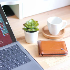 brown leather bifold wallet beside white ceramic mug on macbook pro