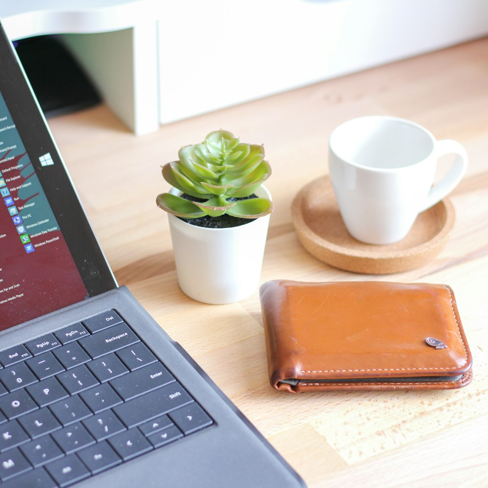 brown leather bifold wallet beside white ceramic mug on macbook pro