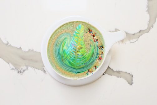 A cup of coffee with colorful latte art featuring vibrant green and blue swirls in the shape of a leaf, accented with multicolored sprinkles on the foam. The cup is placed on a white surface with gray marble-like patterns.
