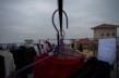 A balcony scene with clothes hanging on a line. Various garments, including a red piece of clothing, are clipped onto hangers. The background is slightly out of focus, showing a cloudy sky and rooftops. The perspective is from the middle of the clothesline, looking across multiple hangers.