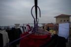 A balcony scene with clothes hanging on a line. Various garments, including a red piece of clothing, are clipped onto hangers. The background is slightly out of focus, showing a cloudy sky and rooftops. The perspective is from the middle of the clothesline, looking across multiple hangers.