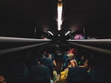 The dimly lit interior of a bus with passengers seated. Personal belongings and bags are stored in the overhead compartments. The focus is on the passengers who are engaged in different activities, possibly traveling at night.