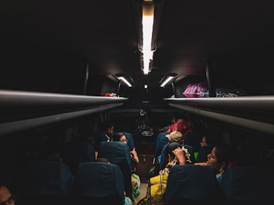 The dimly lit interior of a bus with passengers seated. Personal belongings and bags are stored in the overhead compartments. The focus is on the passengers who are engaged in different activities, possibly traveling at night.
