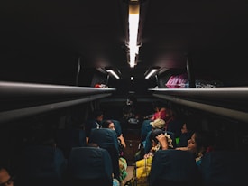 The dimly lit interior of a bus with passengers seated. Personal belongings and bags are stored in the overhead compartments. The focus is on the passengers who are engaged in different activities, possibly traveling at night.