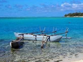 A traditional outrigger canoe is gently resting on the clear, shallow waters of a tropical beach. The ocean stretches out in various shades of blue and turquoise, meeting the horizon where a small island covered with lush greenery is visible. The sky is dotted with fluffy white clouds.