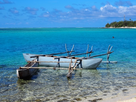 A traditional outrigger canoe is gently resting on the clear, shallow waters of a tropical beach. The ocean stretches out in various shades of blue and turquoise, meeting the horizon where a small island covered with lush greenery is visible. The sky is dotted with fluffy white clouds.