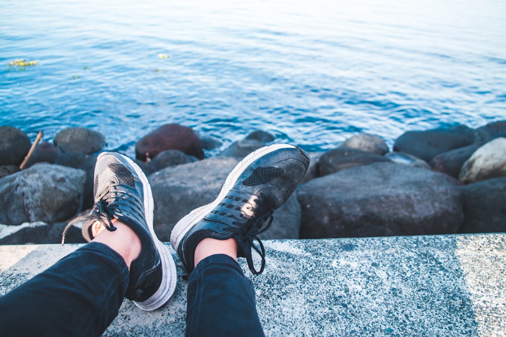 person in black pants and black and white sneakers sitting on rock near body of water