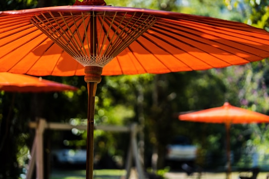 Large orange parasols stand against a blurred natural background, casting patterns of light and shadow. The focus on the parasol in the foreground highlights its intricate framework and vibrant color, while similar structures are subtly visible in the distance.
