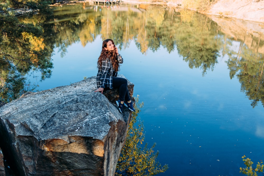 woman in black jacket sitting on rock near lake during daytime