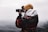A person wearing a patterned jacket and a mustard yellow beanie is taking a photograph with a DSLR camera. The background is a misty, overcast landscape, suggesting a cold, serene environment.