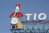 A rooftop billboard against a clear blue sky features a stylized bottle dressed in a red jacket and hat, holding a guitar. The bottle advertises 'Gonzalez Byass Jerez' with the text 'Tio Pepe' on the label. Large letters spell out 'TIO SOL DE ANDA'.