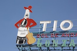 A rooftop billboard against a clear blue sky features a stylized bottle dressed in a red jacket and hat, holding a guitar. The bottle advertises 'Gonzalez Byass Jerez' with the text 'Tio Pepe' on the label. Large letters spell out 'TIO SOL DE ANDA'.