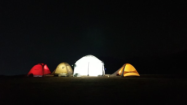 A group of tents set up on a field at night with one brightly illuminated from within. The tents are of various colors, including red, beige, and orange, and are pitched on a dark, expansive ground under a black sky.