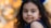A young girl with long, curly black hair and a gentle expression looks towards the camera. She wears a dark blue outfit and has vibrant blue feather-like decorations in her hair. The background is softly blurred with warm, autumnal orange and yellow bokeh lights.