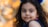 A young girl with long, curly black hair and a gentle expression looks towards the camera. She wears a dark blue outfit and has vibrant blue feather-like decorations in her hair. The background is softly blurred with warm, autumnal orange and yellow bokeh lights.