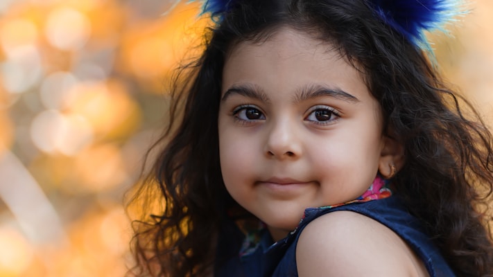 A young girl with long, curly black hair and a gentle expression looks towards the camera. She wears a dark blue outfit and has vibrant blue feather-like decorations in her hair. The background is softly blurred with warm, autumnal orange and yellow bokeh lights.