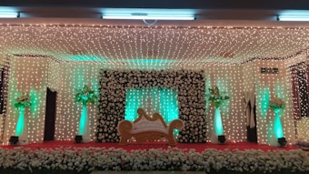 A decorative stage setup features a central ornate sofa with a golden frame, flanked by columns adorned with green lights and floral arrangements. The backdrop is covered in a curtain of small, twinkling lights and a large frame of white roses. A carpet of flowers lines the front of the stage.