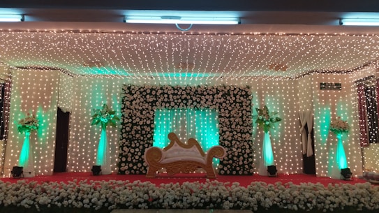 A decorative stage setup features a central ornate sofa with a golden frame, flanked by columns adorned with green lights and floral arrangements. The backdrop is covered in a curtain of small, twinkling lights and a large frame of white roses. A carpet of flowers lines the front of the stage.