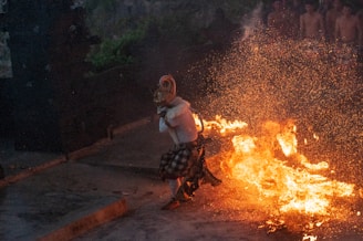 A performer in traditional attire dances amid blazing flames, creating a striking display of fire and movement. The scene appears intense and dynamic, with sparks flying through the air. A group of shirtless individuals is faintly visible in the background, suggesting a ceremonial or cultural event.