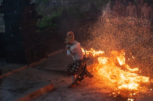 A performer in traditional attire dances amid blazing flames, creating a striking display of fire and movement. The scene appears intense and dynamic, with sparks flying through the air. A group of shirtless individuals is faintly visible in the background, suggesting a ceremonial or cultural event.