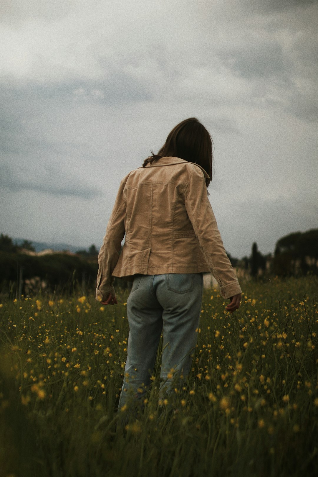 woman in brown jacket and blue denim jeans standing on yellow flower field during daytime