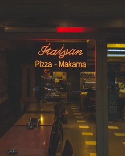A dimly lit restaurant dining area featuring a warm, glowing neon sign reading 'Italyan Pizza - Makarna', surrounded by wooden tables arranged in a row. Chairs are neatly tucked under the tables, and the interior extends into the background with shelves and a counter visible. The ambiance suggests a cozy, intimate dining experience.
