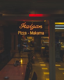 A dimly lit restaurant dining area featuring a warm, glowing neon sign reading 'Italyan Pizza - Makarna', surrounded by wooden tables arranged in a row. Chairs are neatly tucked under the tables, and the interior extends into the background with shelves and a counter visible. The ambiance suggests a cozy, intimate dining experience.