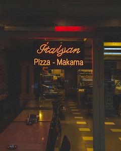 A dimly lit restaurant dining area featuring a warm, glowing neon sign reading 'Italyan Pizza - Makarna', surrounded by wooden tables arranged in a row. Chairs are neatly tucked under the tables, and the interior extends into the background with shelves and a counter visible. The ambiance suggests a cozy, intimate dining experience.
