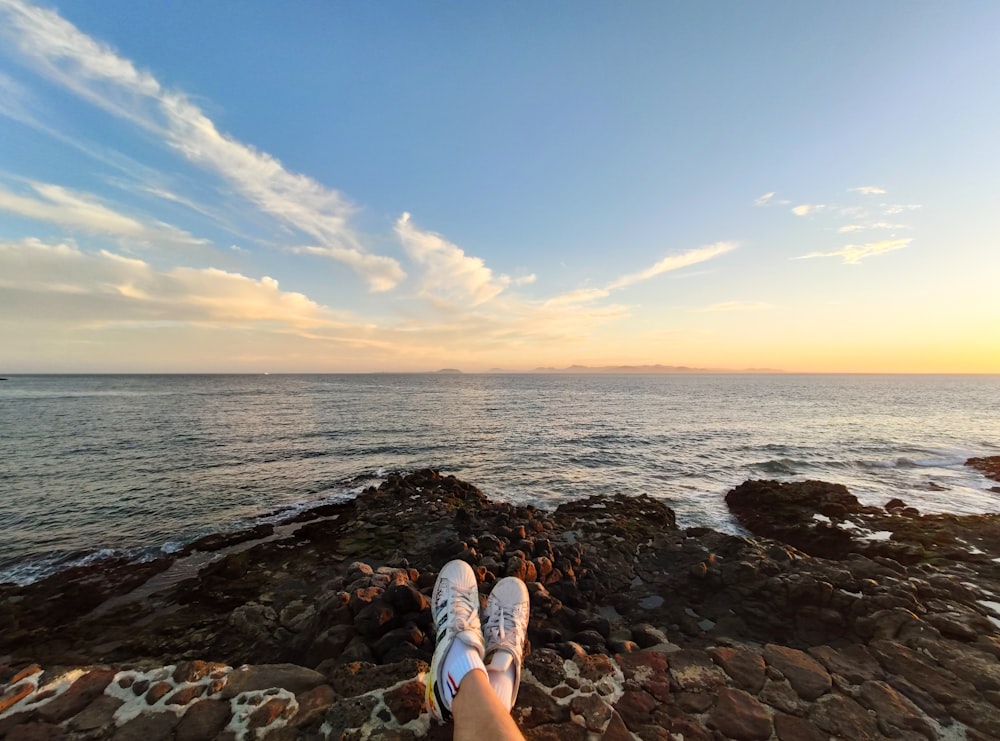 personne en baskets bleues et blanches assise sur un rivage rocheux pendant la journée