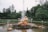 A statue of a man with a trident and a sea creature, surrounded by a fountain with four turtle sculptures. The scene is set in a lush green park with trees and bushes. People can be seen walking and sitting on benches in the background.