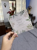 A hand is holding a card with foliage designs and an inspirational quote by A. A. Milne. The background shows a room with a bed covered in gray bedding, a wall clock, red flowers in a vase, and a globe. Natural light is coming in through a window with sheer curtains.