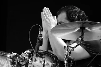 A friendly portrait of Andu Hiriart with a camera and drum set in the background.