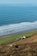 A serene coastal landscape with gentle waves rolling onto a sandy shoreline. A small, isolated farmhouse with several outbuildings sits on lush green fields near the coast. The scene is bordered by a gently sloping hill covered in brown and green vegetation.