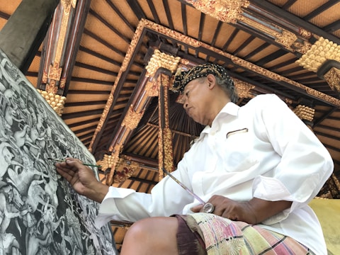 A person wearing a traditional hat and a white shirt is focused on painting a detailed black and white artwork with intricate designs. The background features an ornate ceiling with wooden beams and gold embellishments, reflecting traditional architectural elements.