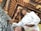 A person wearing a traditional hat and a white shirt is focused on painting a detailed black and white artwork with intricate designs. The background features an ornate ceiling with wooden beams and gold embellishments, reflecting traditional architectural elements.