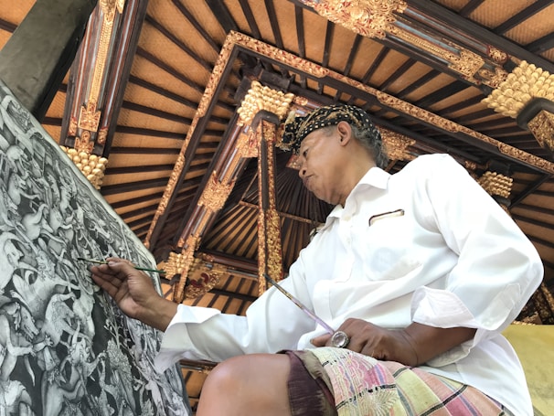 A person wearing a traditional hat and a white shirt is focused on painting a detailed black and white artwork with intricate designs. The background features an ornate ceiling with wooden beams and gold embellishments, reflecting traditional architectural elements.