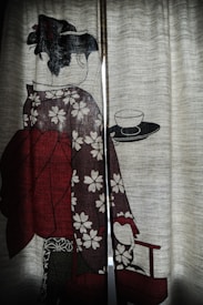 A traditional Japanese illustration is depicted on a curtain, featuring a woman in a kimono adorned with floral patterns. She is holding a tray with a cup and saucer. The fabric contains muted tones and allows light to pass through, creating a slightly translucent effect.