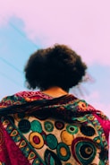A person is viewed from the back, showcasing a vibrant and intricately patterned fabric draped over their shoulders. The design features circular motifs in rich colors, including gold, turquoise, and red. The background consists of a clear sky with faint cloud streaks.