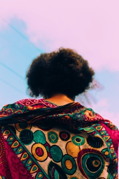 A person is viewed from the back, showcasing a vibrant and intricately patterned fabric draped over their shoulders. The design features circular motifs in rich colors, including gold, turquoise, and red. The background consists of a clear sky with faint cloud streaks.