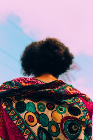 A person is viewed from the back, showcasing a vibrant and intricately patterned fabric draped over their shoulders. The design features circular motifs in rich colors, including gold, turquoise, and red. The background consists of a clear sky with faint cloud streaks.
