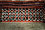 Rows of loudspeakers are mounted on a red wall, each displaying various words, likely indicating language or sound-related themes. The setup exudes a sense of uniformity and organization.