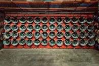 Rows of loudspeakers are mounted on a red wall, each displaying various words, likely indicating language or sound-related themes. The setup exudes a sense of uniformity and organization.