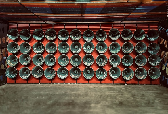 Rows of loudspeakers are mounted on a red wall, each displaying various words, likely indicating language or sound-related themes. The setup exudes a sense of uniformity and organization.