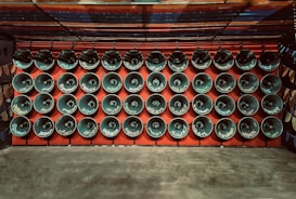 Rows of loudspeakers are mounted on a red wall, each displaying various words, likely indicating language or sound-related themes. The setup exudes a sense of uniformity and organization.