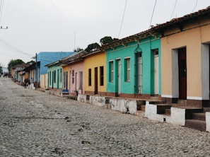A row of colorful, single-story houses lines a cobblestone street. Each house is painted in bright colors such as blue, green, yellow, and peach. The street is quiet with a few people in the distance.